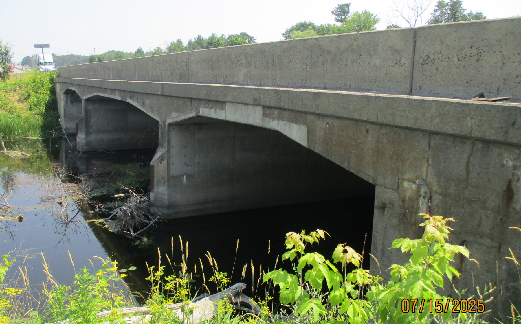 Concrete bridge spanning a shallow river, showing multiple support arches and vegetation along the riverbank.